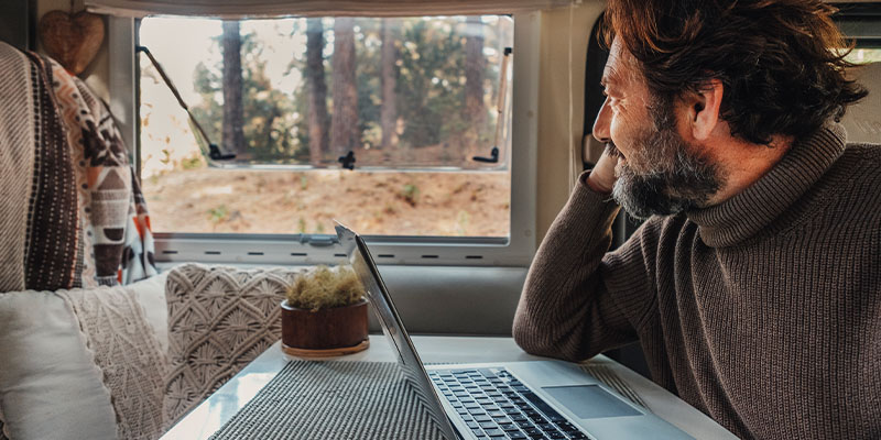 En man sitter och tittar ut genom sitt husvagnsfönster samtidigt som han har en dator framför sig.