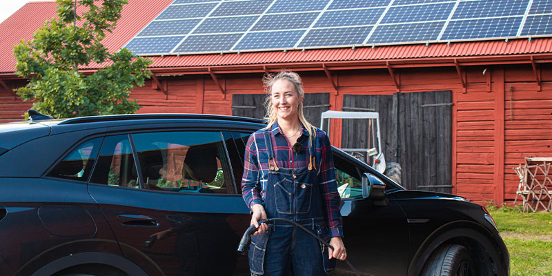 Bonden Emelie står framför sin elbil som laddas med solcellerna på ladugårdstaket.