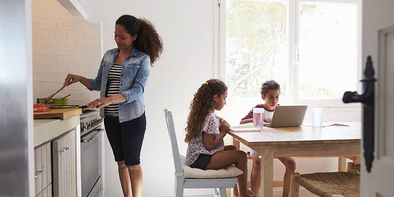 En lägenhet där mamma lagar mat och två barn sitter vid ett bord varav ena skriver på en dator.
