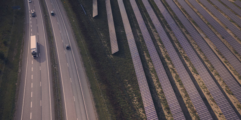 Bilar och en lastbil kör på en väg bredvid en solcellspark med flera rader av solpaneler.