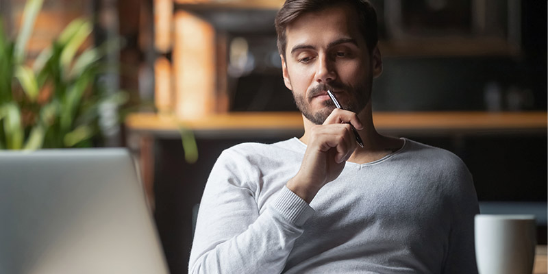 En man sitter och läser på sin dator med en kopp kaffe bredvid sig och funderar.